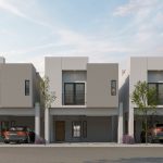 La imagen muestra una fila de casas modernas de dos pisos en un fraccionamiento residencial. Las fachadas de las casas son de estilo minimalista, con líneas limpias y colores neutros, predominantemente en tonos de blanco y gris. Cada casa tiene un garaje en la planta baja, algunos de los cuales están ocupados por automóviles. En el segundo piso, se pueden ver grandes ventanas y balcones pequeños. Hay árboles jóvenes plantados en la acera frente a las casas, y arbustos de lavanda decoran el área cercana a las entradas. El cielo está parcialmente nublado, sugiriendo un día tranquilo y sereno.